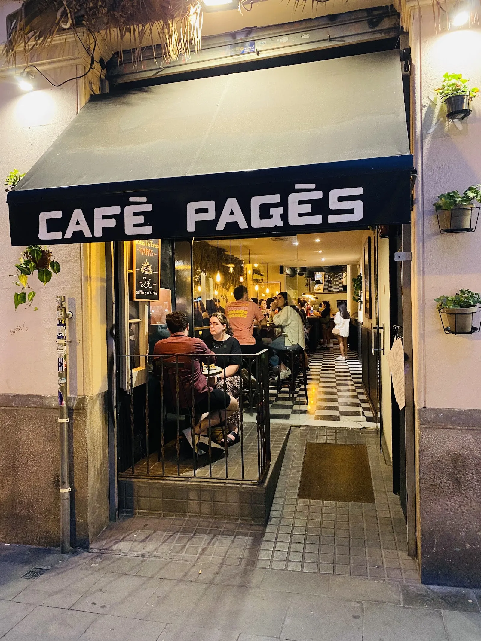 Fachada de Cafè Pagès con su toldo negro, de noche, con gente en las mesas de dentro
